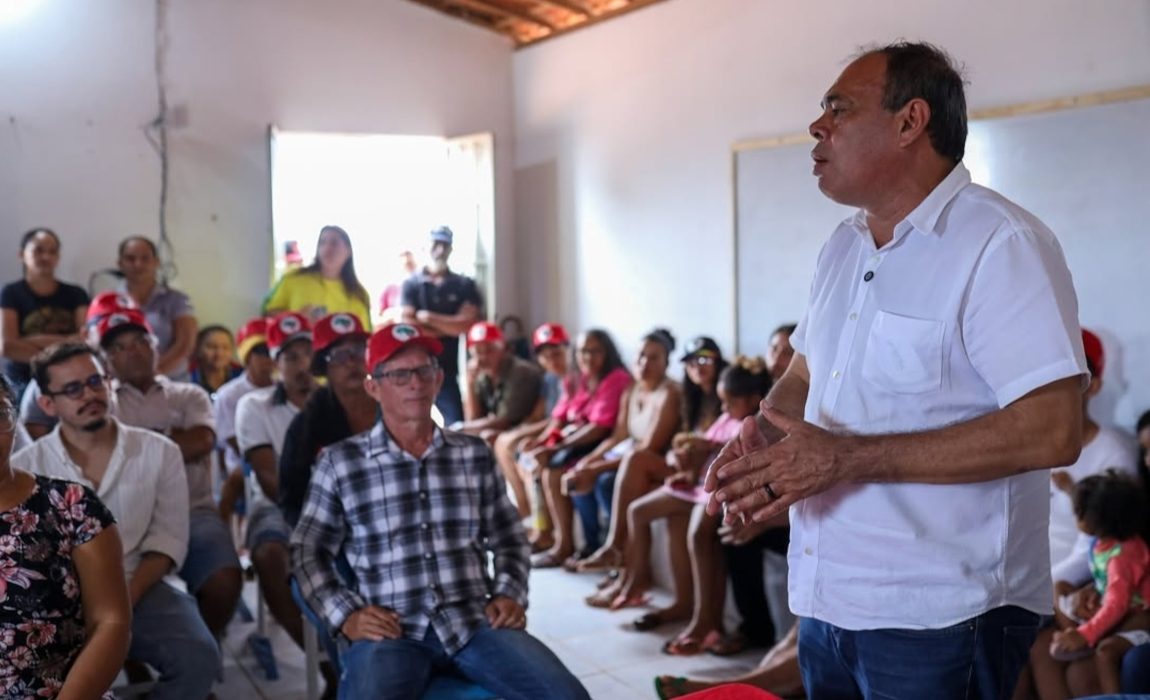 Um homem de camiseta branca em pé fala para várias pessoas sentadas em uma sala grande.