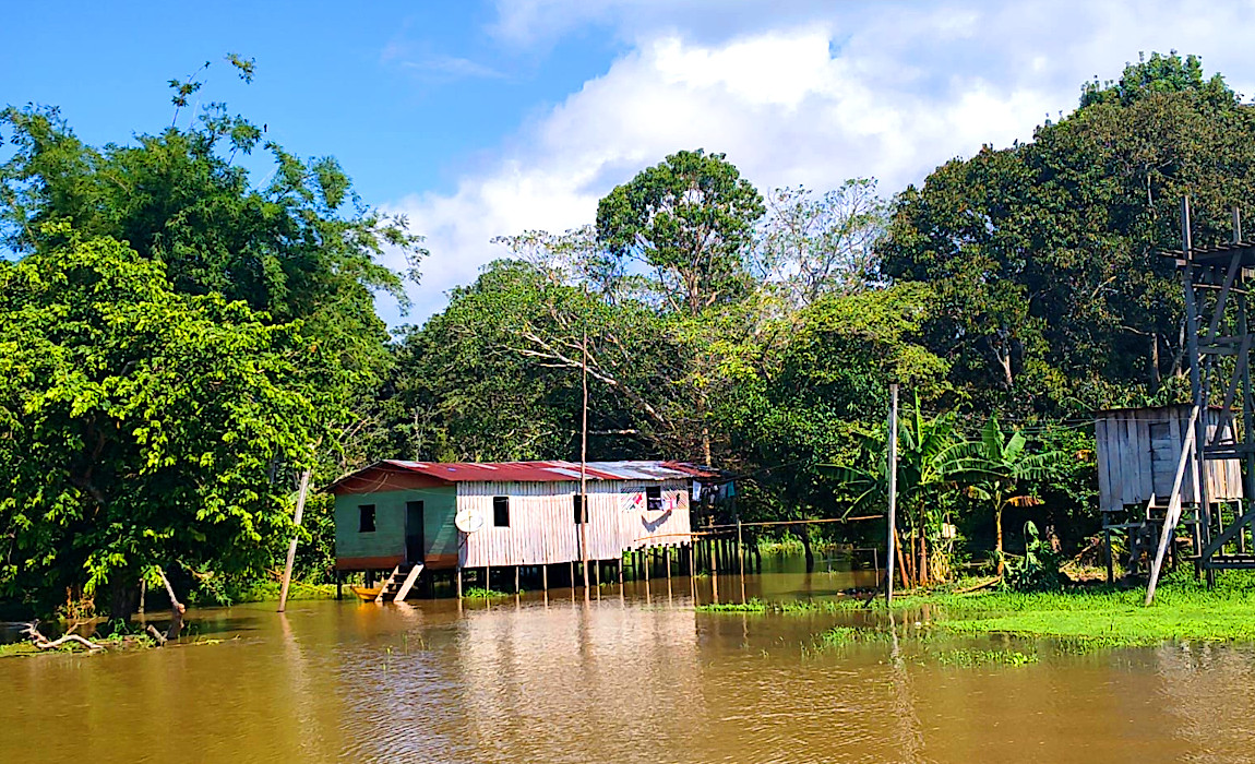 Imagem mostra casa branca com teto roxo em palafitas sobre beira de rio, com mata ao lado e ceu azul acima