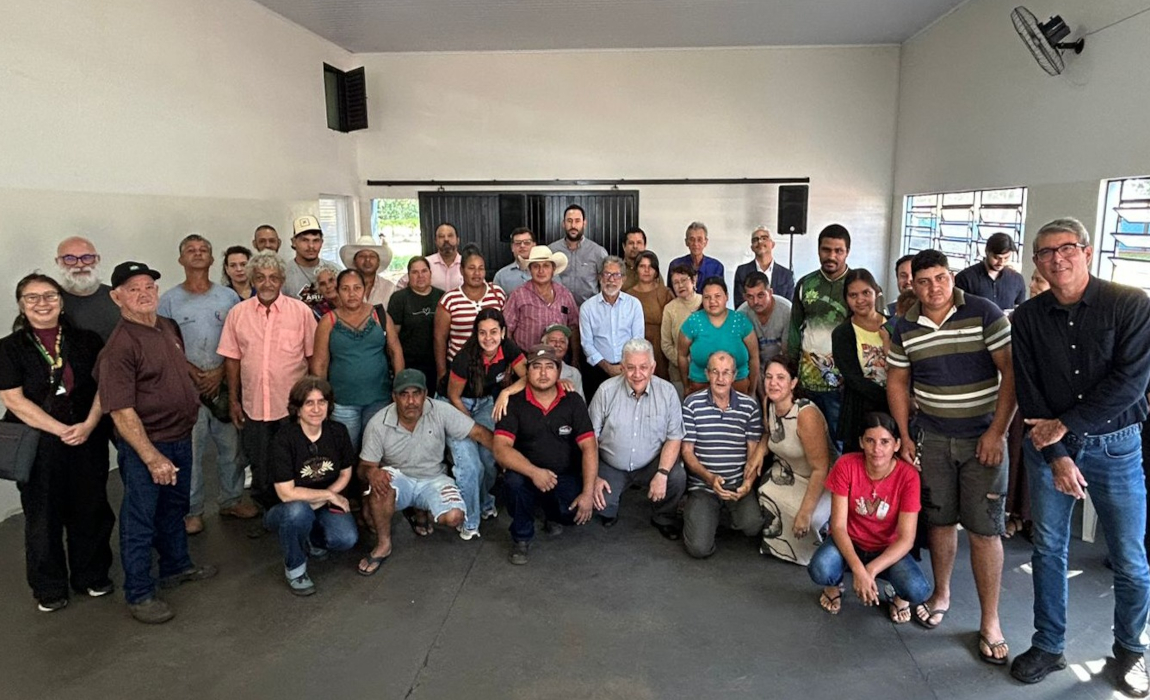 Um grupo numeroso de pessoas, incluindo homens e mulheres de diversas idades, posa para uma foto em um ambiente interno. Alguns estão sentados no chão, enquanto a maioria fica em pé atrás deles.