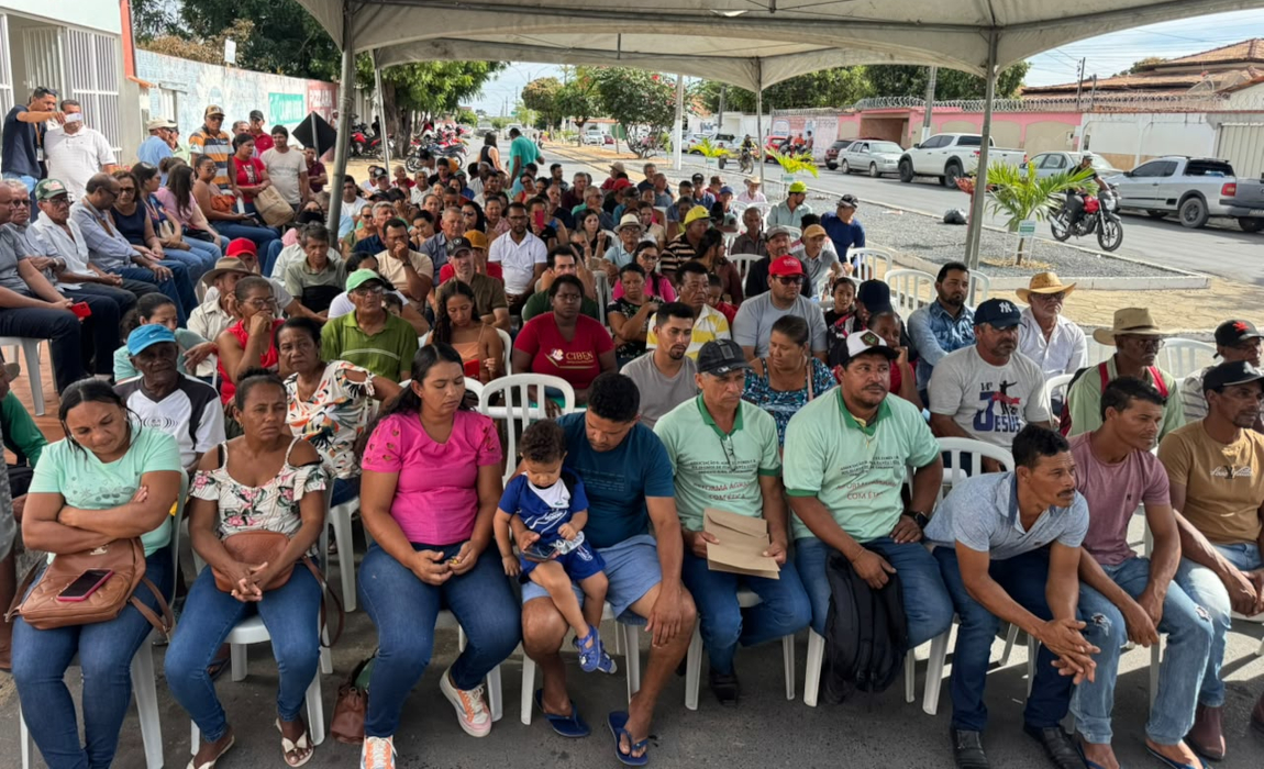 Um grupo grande de pessoas, homens e mulheres, estão sentados em uma espécie de galpão.