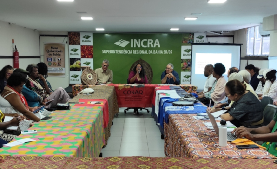 Um grupo de pessoas sentadas em mesas organizadas em forma de U, participando de uma reunião ou evento. Ao fundo, um painel verde exibe a marca do Incra.