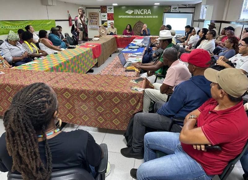 Um grupo de pessoas sentadas em mesas organizadas em forma de U, participando de uma reunião.