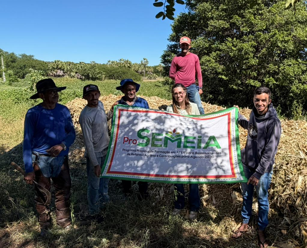Homens e mulheres em pé, segurando um cartaz com o nome Pro-Semeia, em um campo aberto, com uma árvo