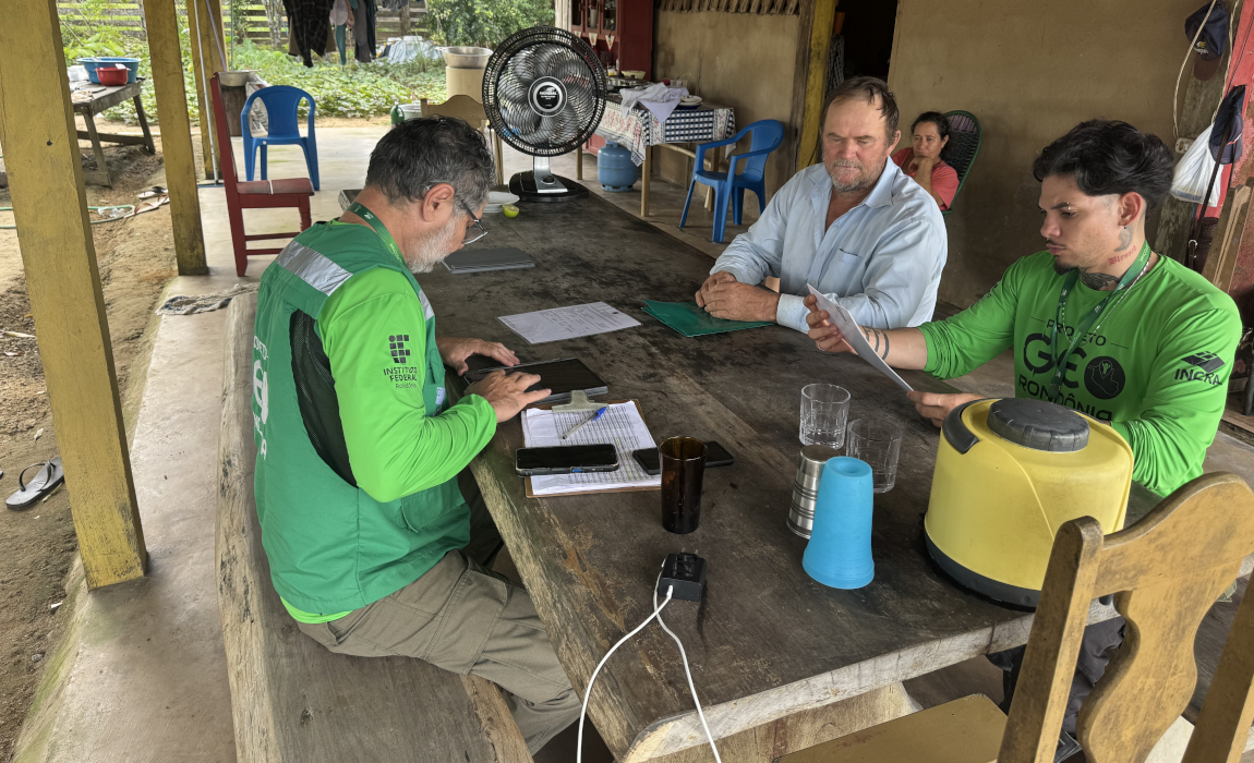 Três homens estão sentados em uma mesa de madeira ao ar livre. Um homem vestindo um colete verde com um logo federal está olhando para uma tela de tablet, enquanto outro homem, vestido com uma camisa azul clara, segura papéis. O terceiro homem está lendo um documento em sua frente.