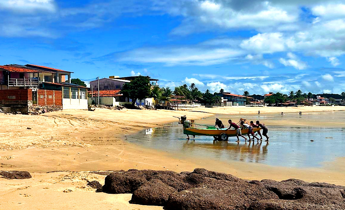 Imagem mostra uma práia de colônia de pescadores, com algums casas ao fundo e homens retirando barco do mar