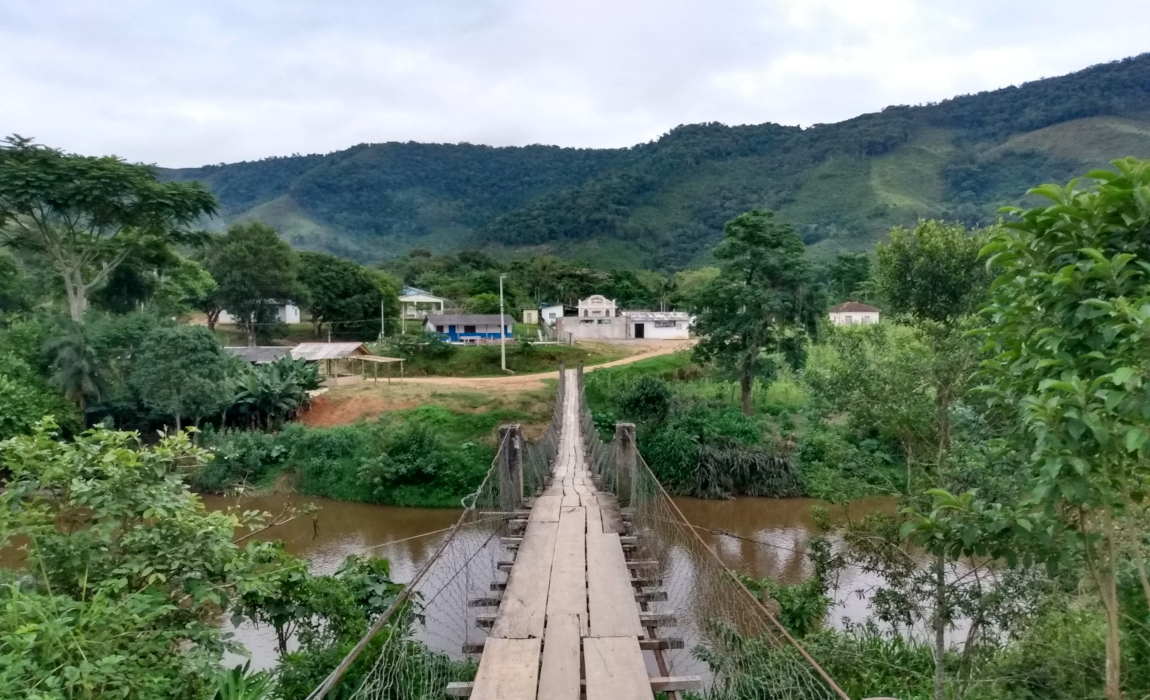 Um córrego com uma ponte de madeira e do outro lado casas e outras construções