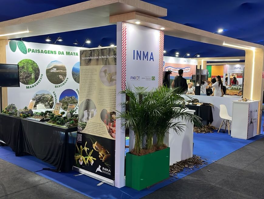 Estande do INMA (Instituto Nacional da Mata Atlântica) na ExpoT&C. O espaço tem estrutura em madeira clara e piso azul. Na lateral esquerda, há um painel com o título "Paisagens da Mata Atlântica, ilustrando ambientes como manguezais, restingas e florestas, além de uma mesa com maquetes.