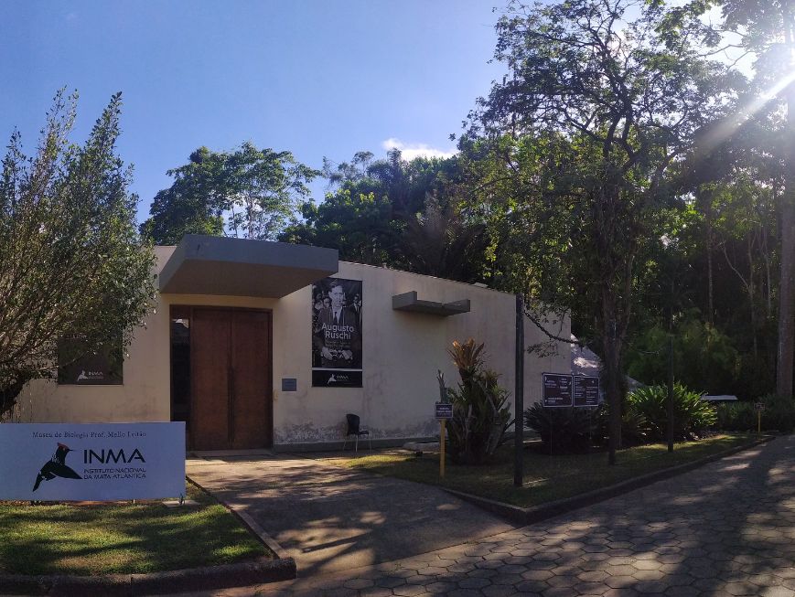 Foto da área externa do Museu de Biologia Professor Mello Leitão, em Santa Teresa (ES). Ao centro, há o auditório com paredes claras, com uma porta de madeira e um painel com a imagem e o nome de Augusto Ruschi. À esquerda, em primeiro plano, há uma placa com a identificação do INMA.