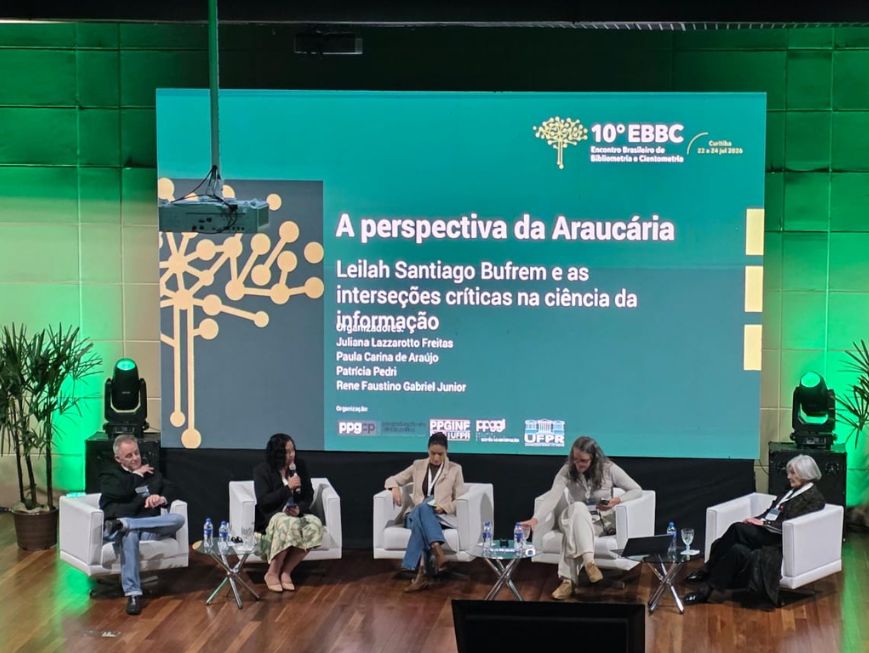 Fotografia de um painel de debate em um auditório. No palco, cinco pessoas estão sentadas em poltronas brancas dispostas em semicírculo, com pequenas mesas de vidro entre elas.
