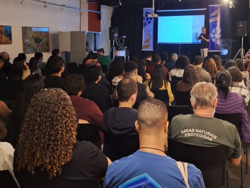 Fotografia de palestra realizada no Auditório Augusto Ruschi. O público está sentado em cadeiras, de costas para a câmera, acompanhando a apresentação. Ao fundo, uma pessoa está em pé, ao lado de uma tela de projeção com conteúdo apresentado em slides.