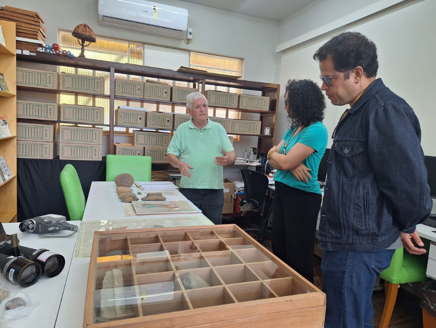 Imagem mostra três pessoas em uma sala de arquivo e pesquisa, observando materiais arqueológicos dispostos sobre uma mesa