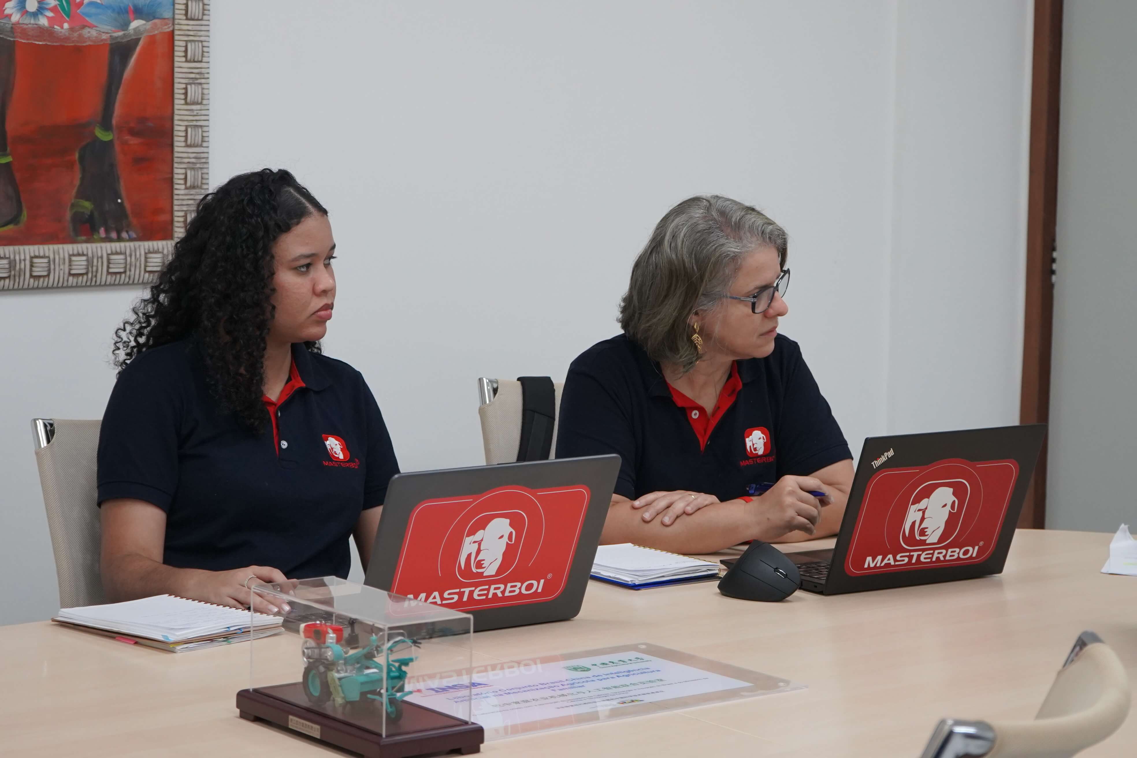 Mulheres com farda da empresa Masterboi diante de computadores na mesa de reuniões