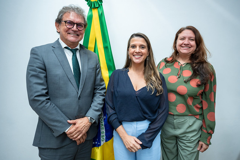 As três pessoas, após a reunião pousam para uma foto atrás da bandeiro do Brasil.