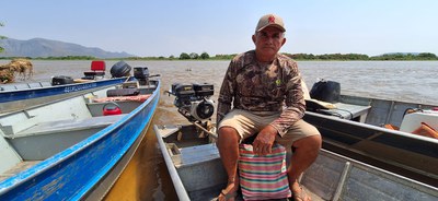 José de Oliveira Amorim, pescador e piloteiro está sentado em seu barco. Há outras embarcações ao redor e o Rio Paraguai ao fundo.jpg