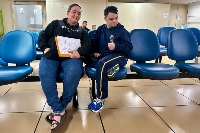 Descrição da imagem: Deise Mara e João Vitor Dahmer sentados em cadeiras azuis na uma sala de espera da Agência da Previdência Social. Foto: Marcos Varjão (ACS SR SUL)