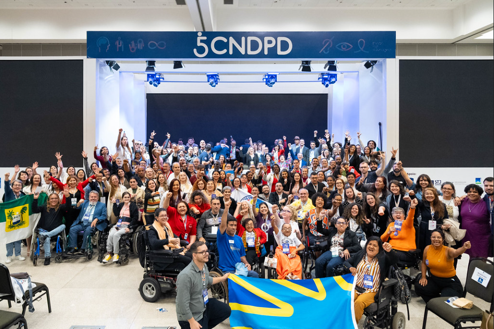 A imagem mostra um grande grupo de pessoas reunidas na 5ª Conferência Nacional dos Direitos da Pessoa Com Deficiência. Elas estão posando para uma foto em frente a um palco com um letreiro que diz "5 CNDPD". Muitas pessoas estão sorrindo e levantando os braços em sinal de celebração. Há uma diversidade de participantes, incluindo pessoas em cadeiras de rodas. Algumas pessoas estão segurando bandeiras, uma delas com um desenho de uma palmeira e outra com um padrão azul e amarelo. O ambiente parece ser um espaço interno, possivelmente um auditório ou centro de convenções.