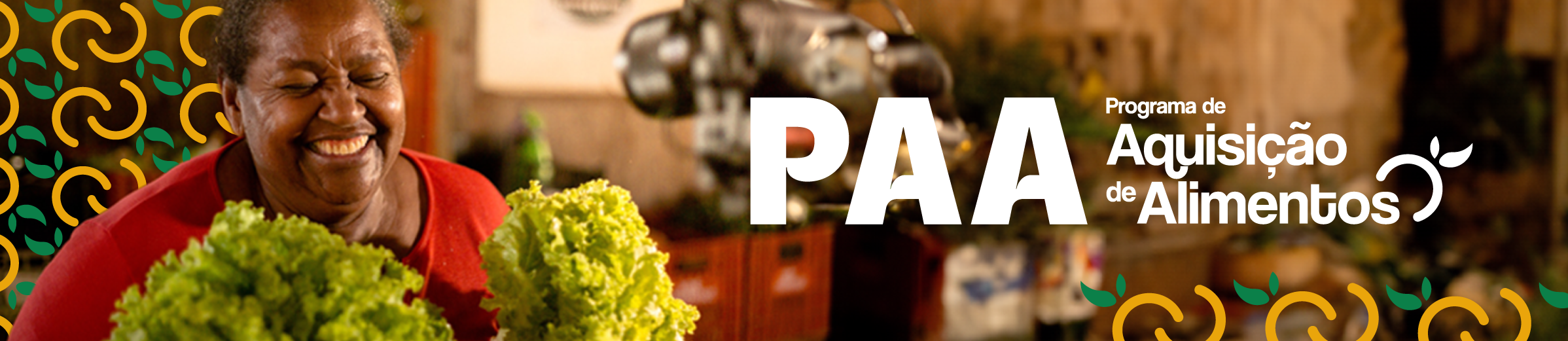 "Mulher sorridente segura dois maços de alface frescos. Ao fundo, elementos gráficos em verde e amarelo decoram a imagem. À direita, aparece o texto ‘PAA – Programa de Aquisição de Alimentos’ em branco."