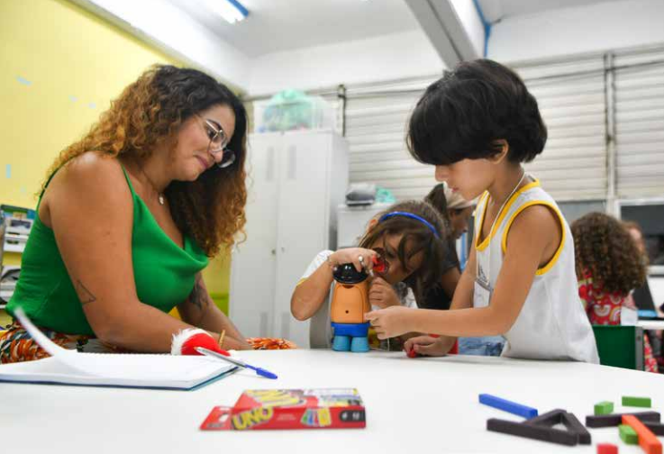 Uma professora observa duas crianças brincando com um boneco educativo em uma sala de aula colorida, enquanto outra criança participa ao fundo. Sobre a mesa há materiais escolares, como lápis e um baralho de cartas UNO.