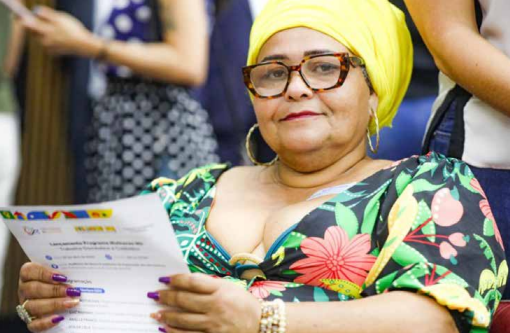 “Mulher com turbante amarelo e óculos segura um documento enquanto participa de um evento público. Ela veste uma roupa colorida com estampas florais e expressa serenidade e orgulho, representando o protagonismo feminino".