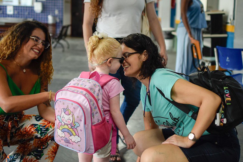 Mulher agachada cumprimenta e sorri para uma menina com mochila rosa nas costas, enquanto outra mulher observa sorrindo ao lado em um ambiente escolar.