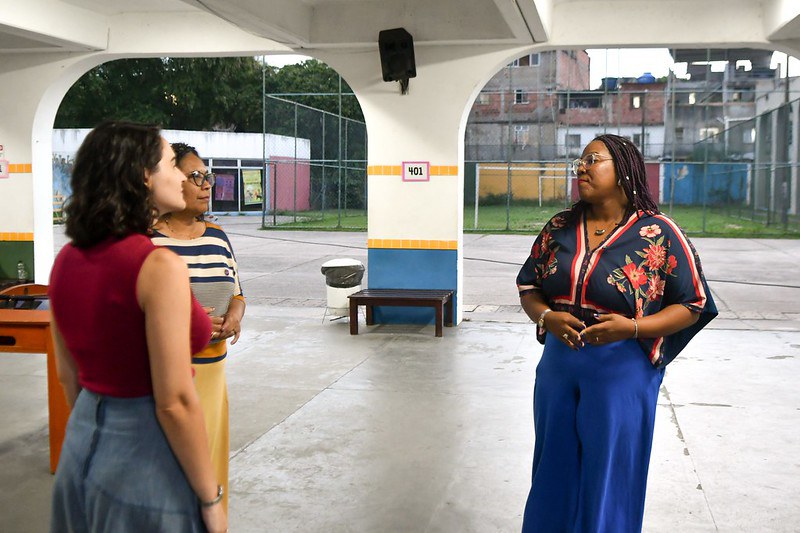 Três mulheres conversam sob uma área coberta de uma escola ou centro comunitário; ao fundo, há um campo esportivo e construções urbanas.