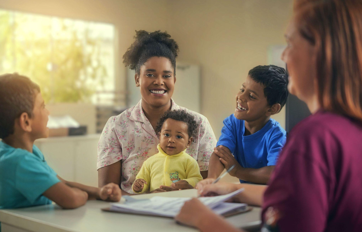 No banner do programa SUAS, há uma imagem onde duas mulheres estão presentes, uma delas está segurando uma caderneta enquanto atende a outra mulher. Ao redor delas, estão três crianças, todas sorrindo. A atmosfera é de alegria e conforto.