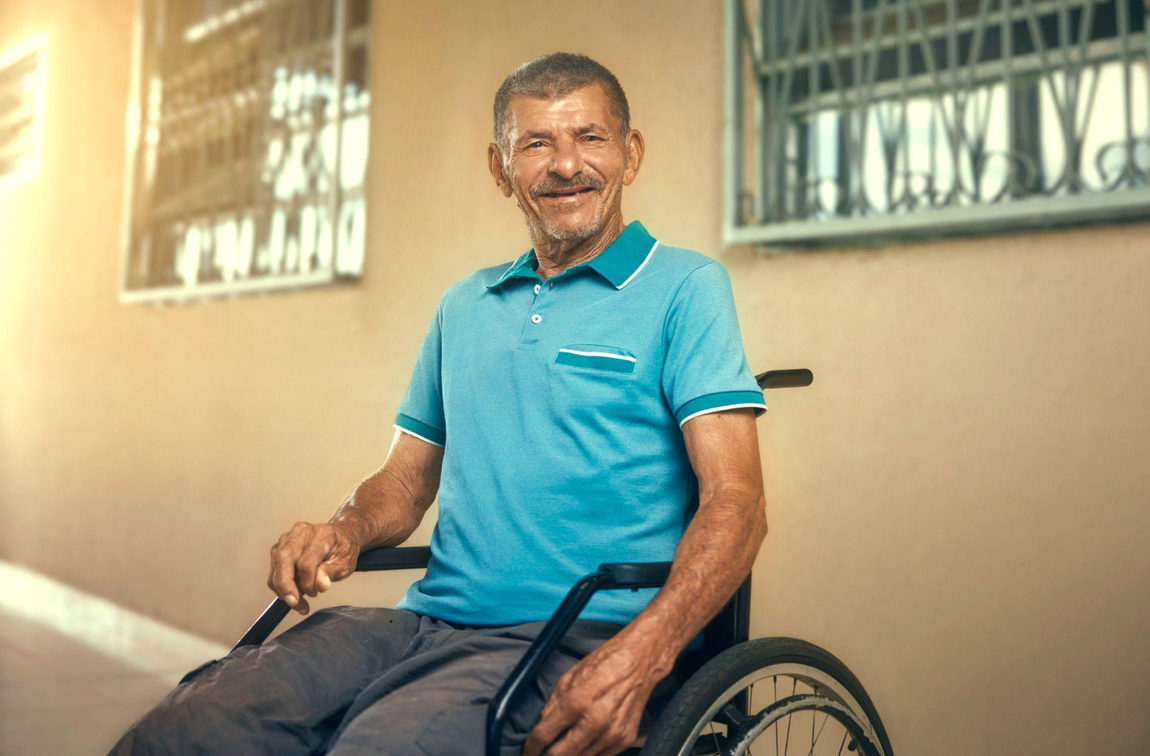Na imagem, há um homem sorridente em uma cadeira de rodas. Ele veste uma blusa azul e calça jeans. Sua expressão é calorosa e acolhedora, transmitindo uma sensação de positividade e confiança.