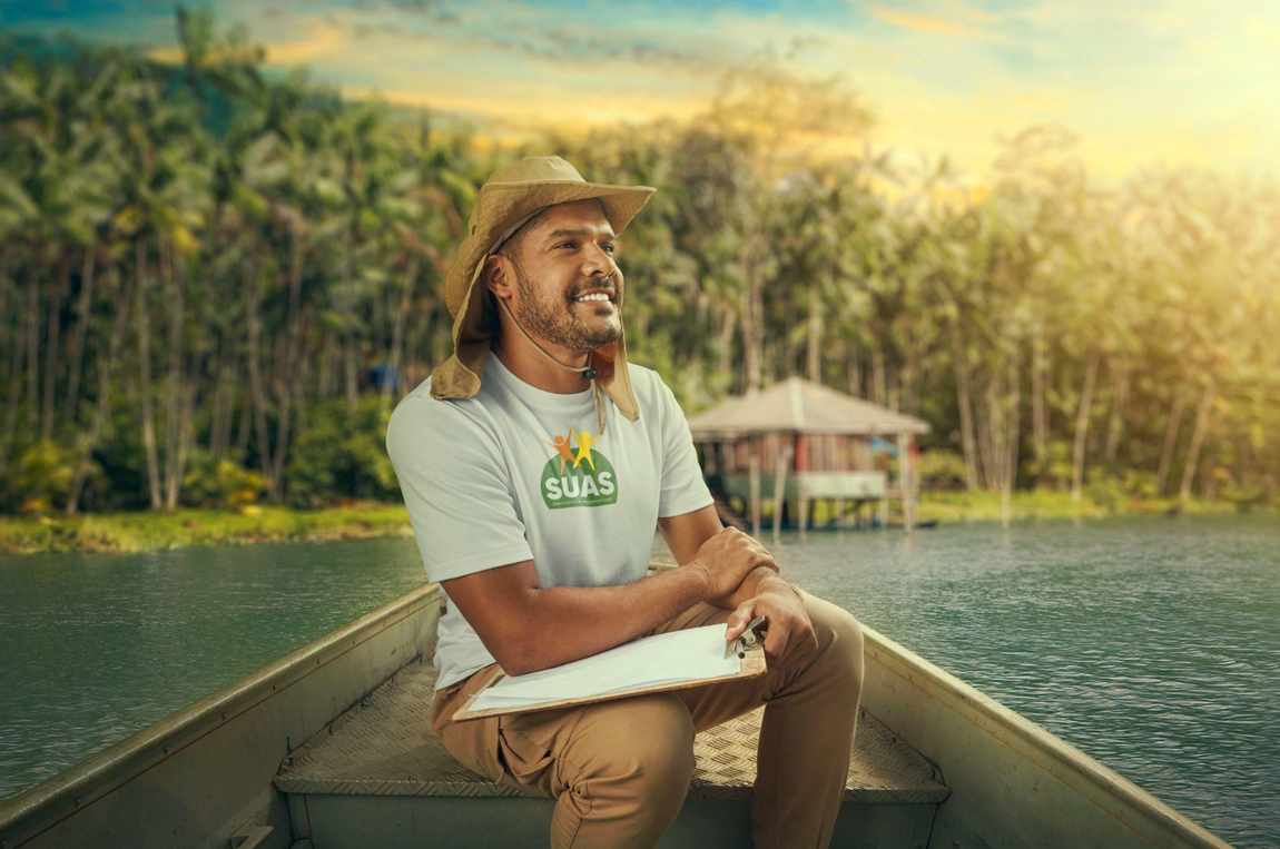 Na imagem, há um homem sorridente usando uma blusa branca do SUAS e segurando uma caderneta. Ele está sentado em um barco, com uma expressão amigável e confiante. O homem veste calça marrom e um chapéu marrom, complementando seu visual enquanto desfruta de sua atividade no barco.