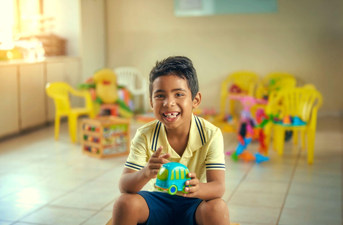 Imagem de uma criança sorridente vestindo uma blusa amarela e um short azul. A criança está segurando um carrinho azul com detalhes verdes e laranjas.