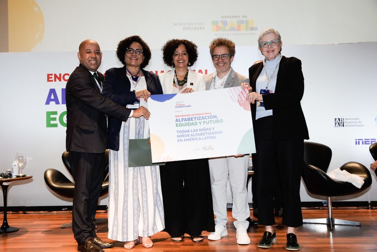 Cinco representantes em palco de evento institucional seguram juntos um painel com o tema “Alfabetização, Equidade e Futuro”, durante encontro sobre alfabetização na América Latina, com logotipos oficiais ao fundo.