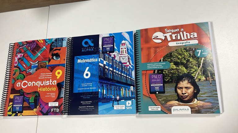 Três livros didáticos do PNLD expostos sobre mesa, incluindo títulos de História, Matemática e Geografia do ensino fundamental, com identificação do programa federal de distribuição de materiais escolares.