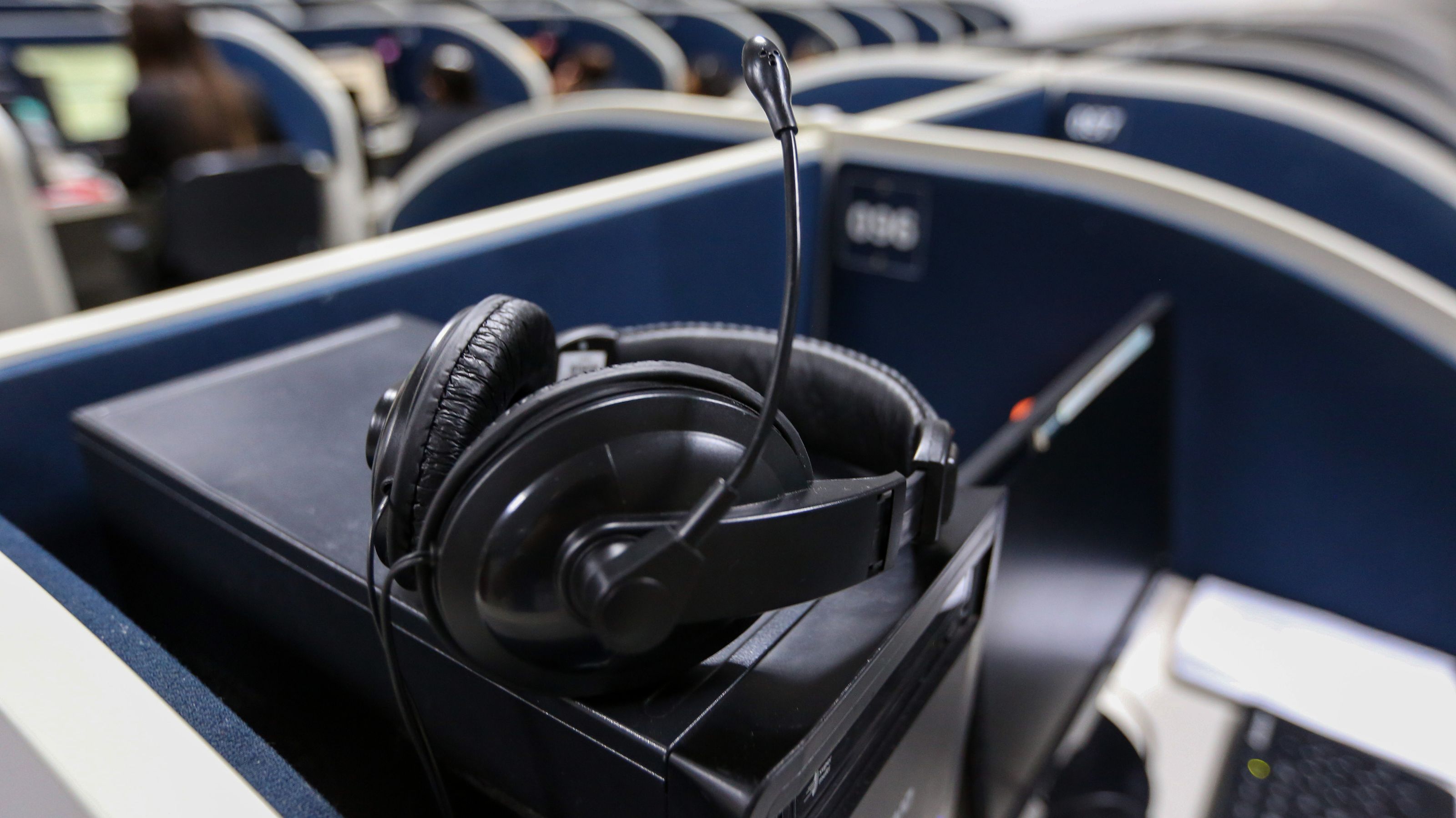 Headset sobre estação de atendimento em central telefônica, com cabines e computadores ao fundo.