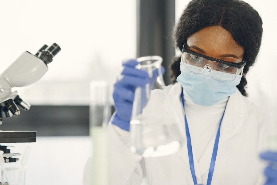 Cientista mulher de jaleco e máscara realizando experimento com microscópio e béquer