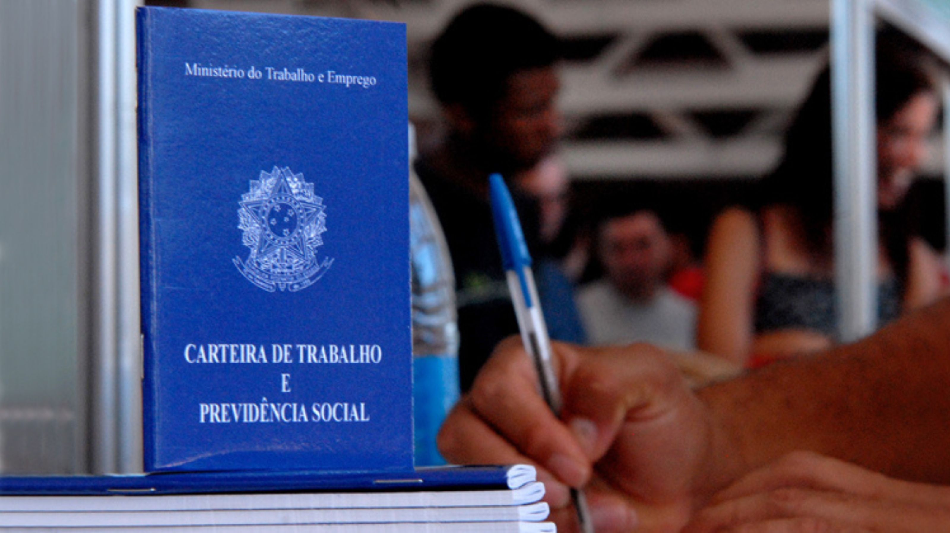 Carteira de Trabalho e Previdência Social (CTPS) azul posicionada em primeiro plano sobre uma bancada. Ao lado, uma pessoa segura uma caneta e escreve em um documento. Ao fundo, outras pessoas aparecem desfocadas em um ambiente de atendimento, sugerindo um contexto relacionado a emprego, contratação ou serviços trabalhistas.