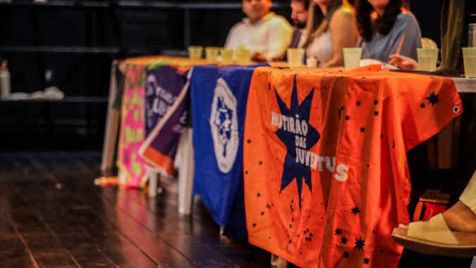 Mesa de plenária com bandeiras de movimentos sociais e participantes ao fundo.