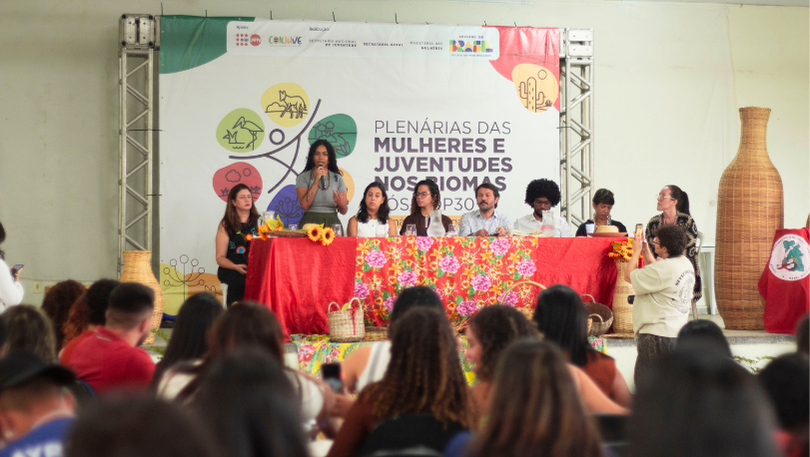 Plenária com mulheres e juventudes em um espaço interno. No palco, uma mesa longa coberta com toalha vermelha e tecido floral reúne oito pessoas sentadas, enquanto uma mulher ao centro, em pé, fala ao microfone. Ao fundo, um banner com o título “Plenárias das Mulheres e Juventudes dos Biomas” e logotipos institucionais. Elementos decorativos como cestos de palha e flores girassóis estão distribuídos sobre a mesa. À frente do palco, o público — majoritariamente feminino — assiste sentado. À direita, uma pessoa registra o momento com o celular. O ambiente é bem iluminado e organizado para um evento de debate ou escuta coletiva.