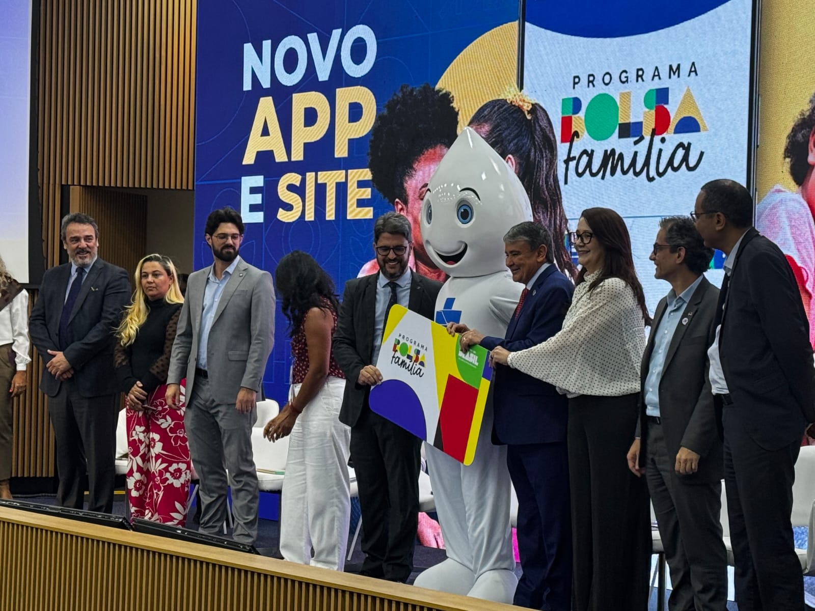Autoridades e participantes posam no palco durante o lançamento do novo aplicativo e site do Programa Bolsa Família. Ao centro, um mascote em formato de gota segura, junto a representantes do governo, uma placa com a identidade visual do programa. Ao fundo, um telão exibe as palavras “Novo app e site” e “Programa Bolsa Família”, com imagens de pessoas. Os participantes estão alinhados lado a lado, vestidos formalmente, em um ambiente institucional.