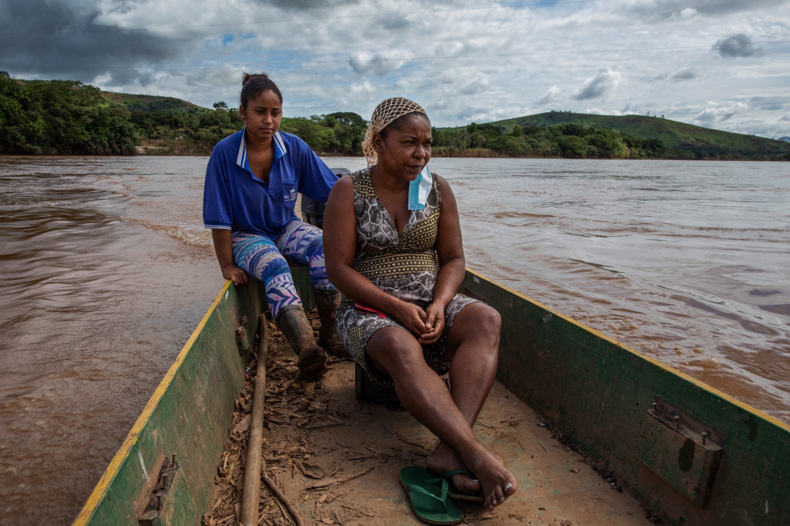 Mulher mais velha e uma jovem estão sentadas em um pequeno barco de madeira, navegando por um rio de águas barrentas. A mulher mais velha está à frente, com expressão séria, usando vestido estampado e um lenço na cabeça; a jovem está atrás, vestindo uma camisa azul e calça estampada. Ao fundo, há vegetação densa e colinas sob um céu nublado.