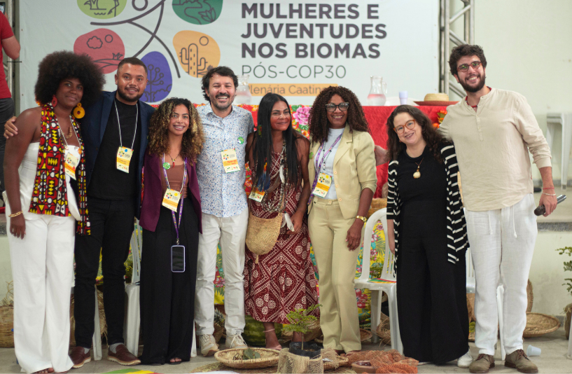 Grupo de oito pessoas posa junto em frente ao banner “Mulheres e Juventudes nos Biomas – Pós-COP30”.