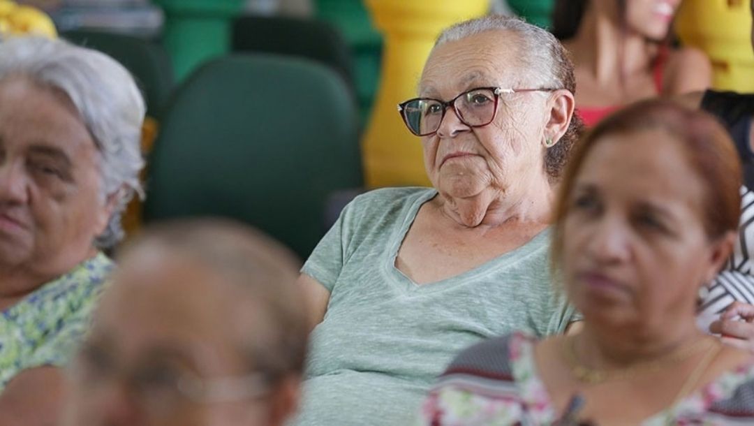 Mulheres idosas sentadas em uma plateia.. Em primeiro plano, uma idosa usa óculos e veste uma blusa azul-esverdeada