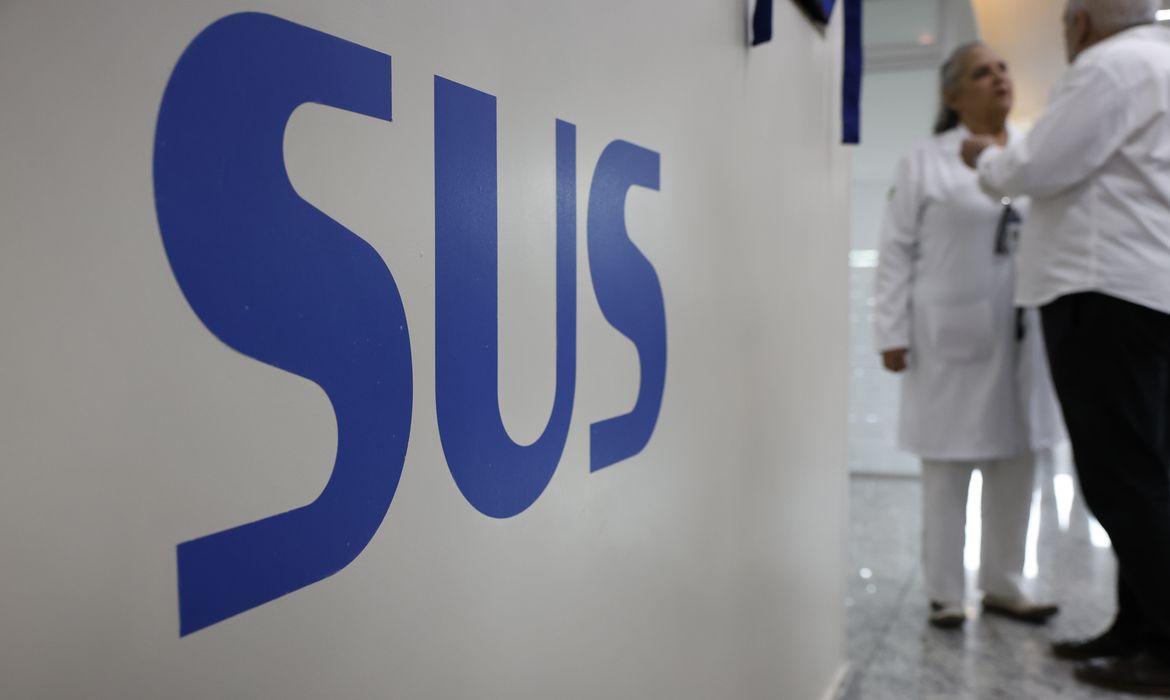 Parede branca em um posto de saúde com a inscrição SUS em azul e em maiúsculas . No corredor, dois profissionais de saúde , uma mulher e um homem estão em pé, conversando.