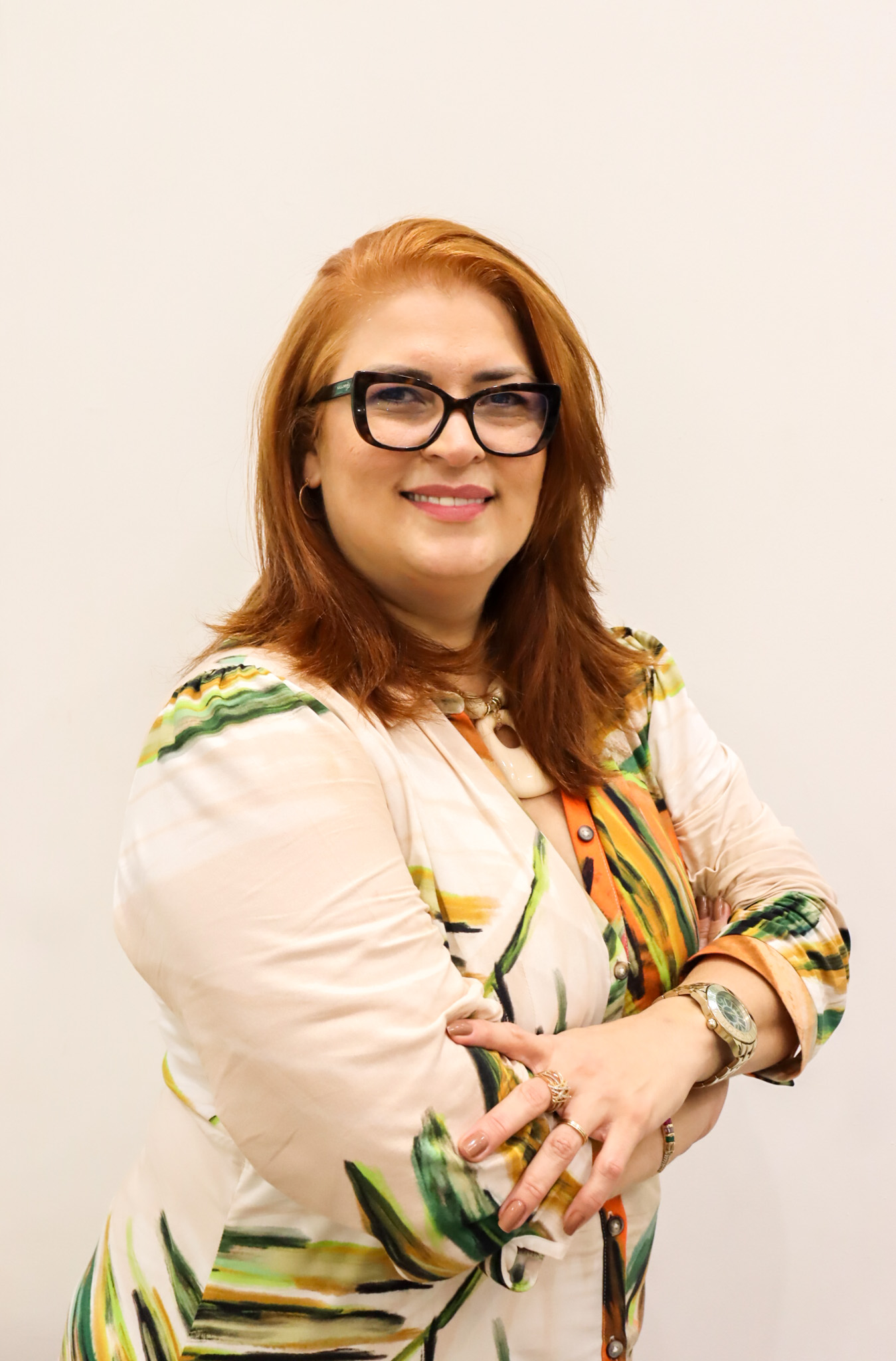 Mulher branca de cabelo ruivo liso usando óculos, camisa com estampa colorida e sorrindo.