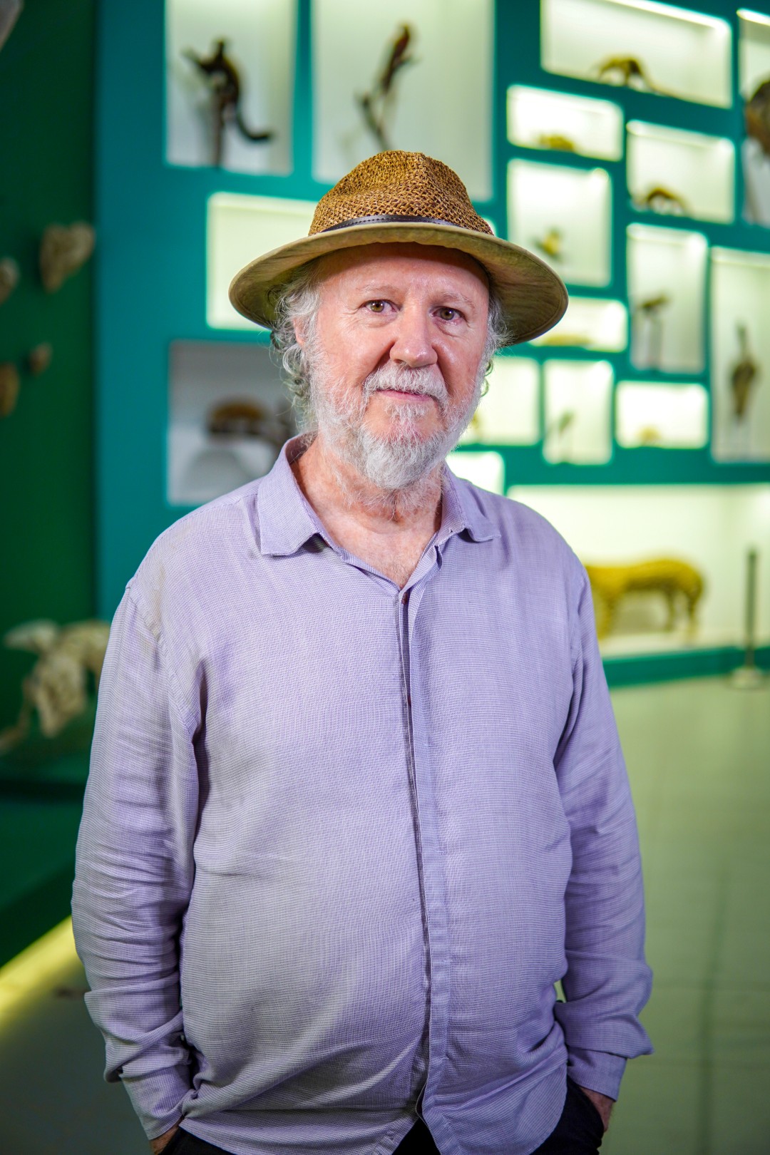Homem branco com barba e cabelo grisalhos usando um chapéu. Ao fundo, vários animais taxidermizados.