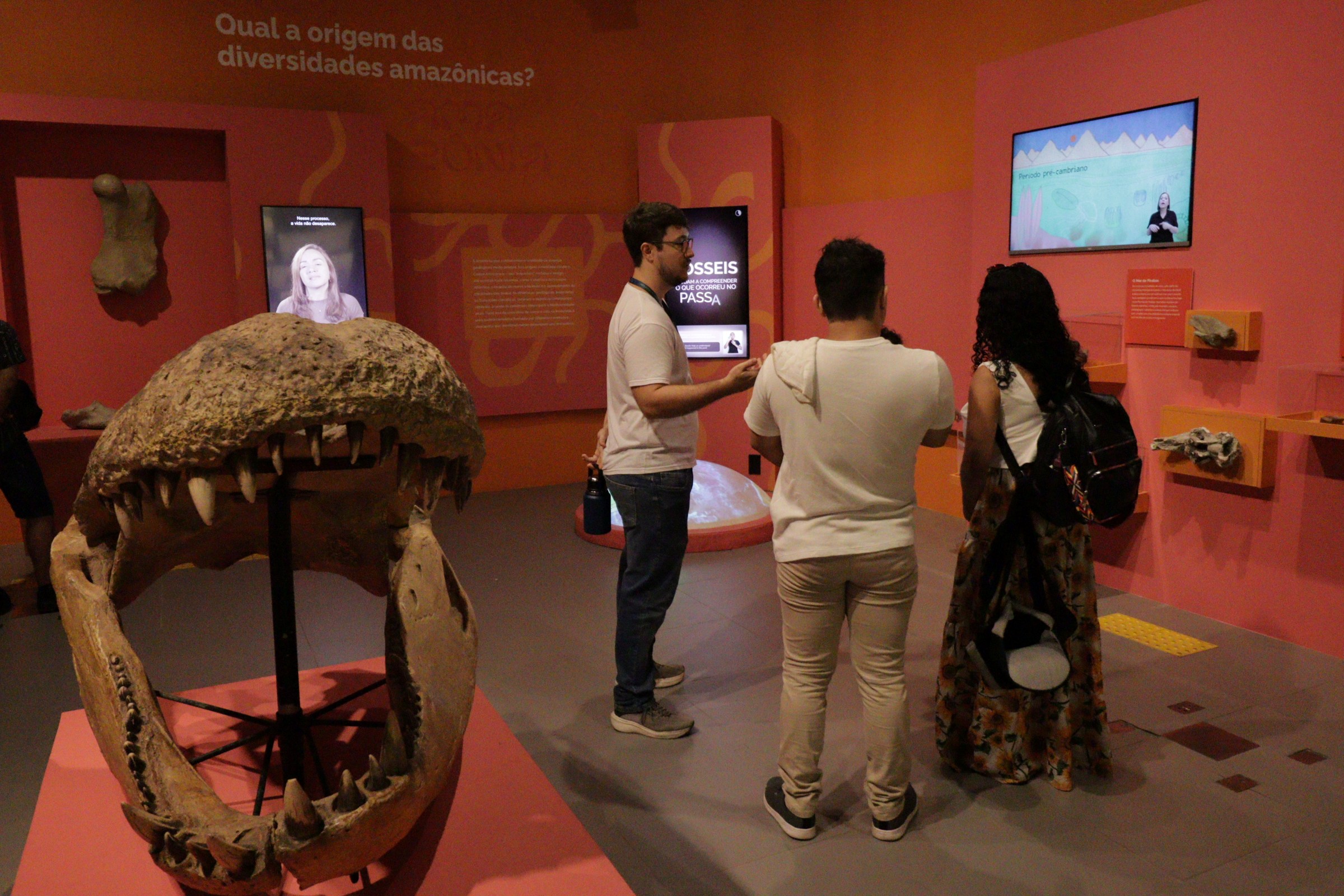 Jovem pesquisador gesticula e interage com casal em ambiente de exposição