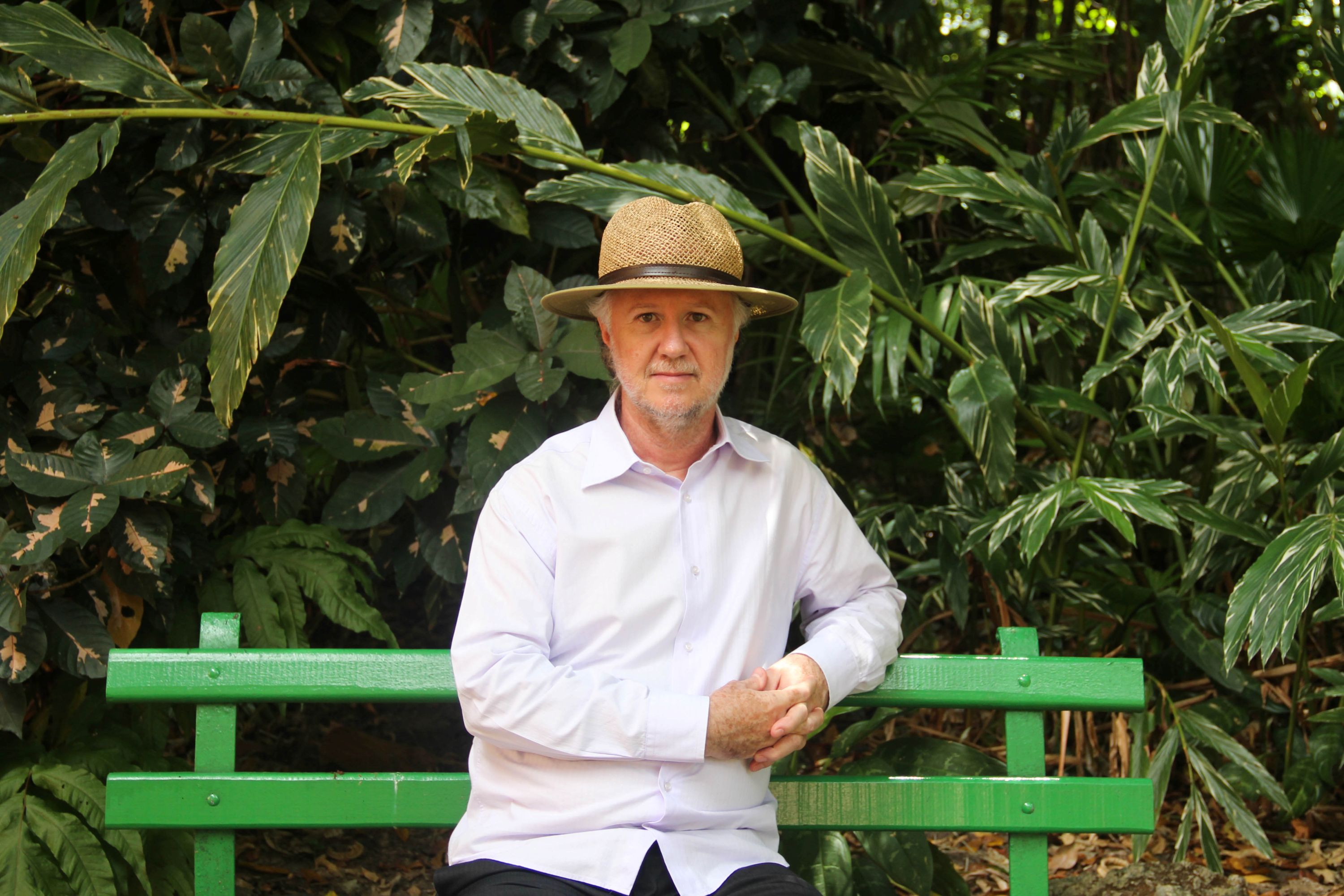 Homem branco, sentado em um banco de madeira pintado de verde, em um ambiente de parque