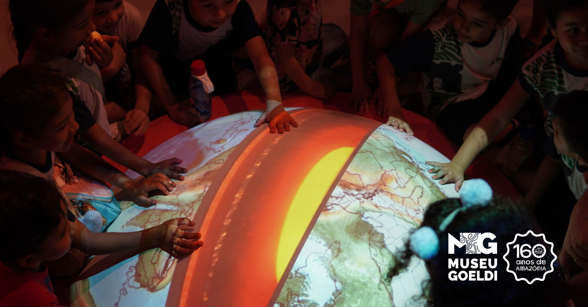 Crianças reunidas ao redor de uma grande esfera que projeta um mapa colorido. Elas tocam e exploram a imagem com as mãos
