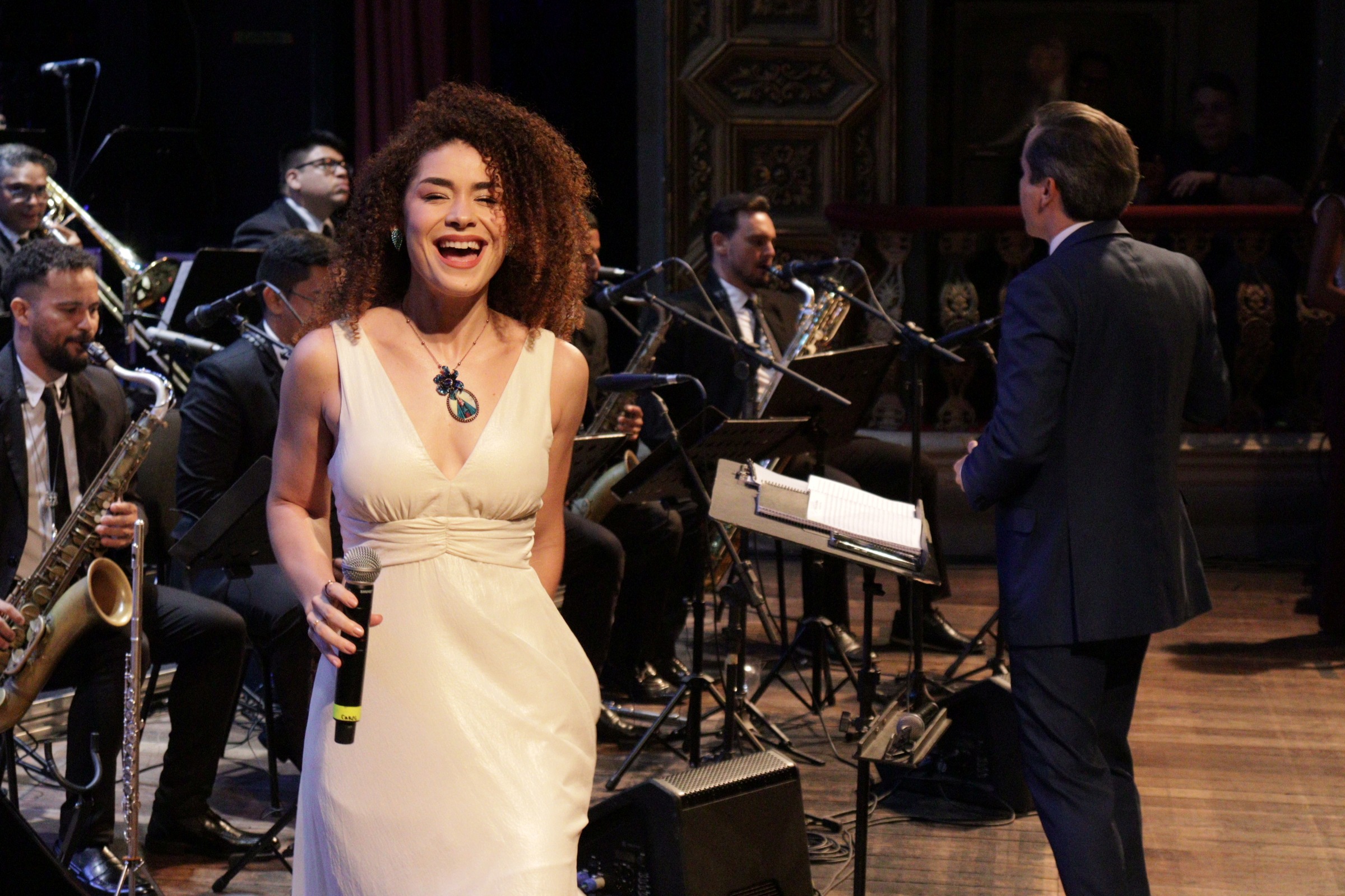 mulher negra com vestido claro sorri em apresentação à frente de orquestra em teatro