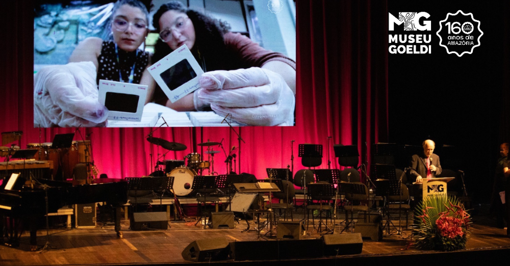 Homem fala em púlpito colocado em palco de teatro enquanto telão exibe imagem de jovens cientistas