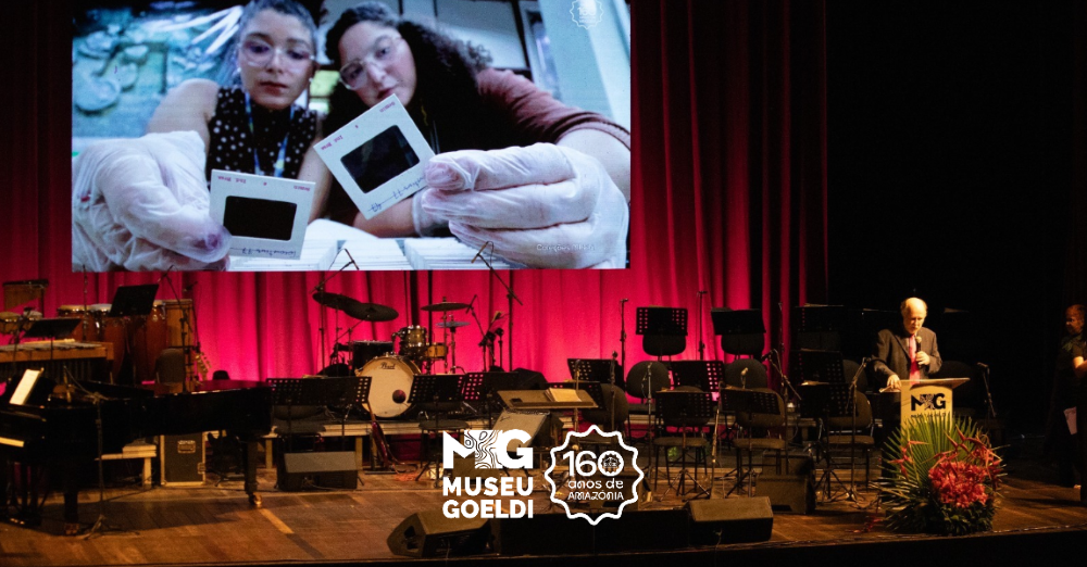Homem fala em púlpito colocado em palco de teatro enquanto telão exibe imagem de jovens cientistas