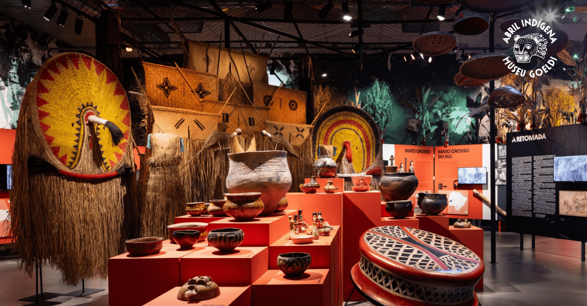 Exposição em museu com artefatos indígenas — cerâmicas, cestos e adornos — organizados sobre bases vermelhas, com grandes peças decorativas ao fundo.